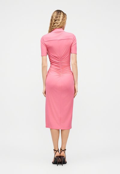 Γυναίκα με κυματιστά μαλλιά που φοράει εφαρμοστή ροζ μίντι φόρεμα με ρίγες στην πλάτη, στέκεται με μαύρα ψηλοτάκουνα σανδάλια μπροστά σε λευκό φόντο.