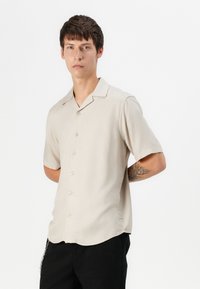 Short-sleeve, beige shirt made of smooth fabric, featuring a pointed collar, six buttons down the front, and a curved hem.
