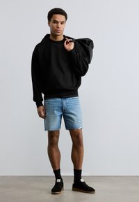 Νεαρός άνδρας που φοράει μαύρο φούτερ, ανοιχτόχρωμα σκισμένα τζιν σορτς, μαύρες ψηλές κάλτσες και αθλητικά παπούτσια, κρατώντας μαύρο σακίδιο στον ώμο του.