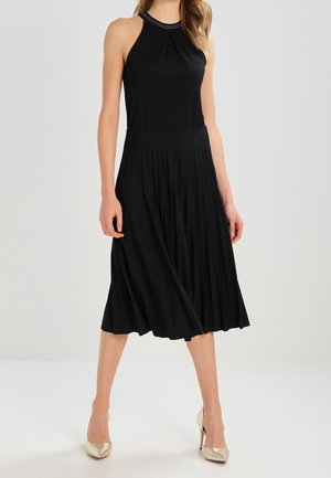 Kobieta ubrana w czarną sukienkę midi bez rękawów z plisowanym dołem i halterkowym dekoltem, zestawioną z beżowymi szpilkami.