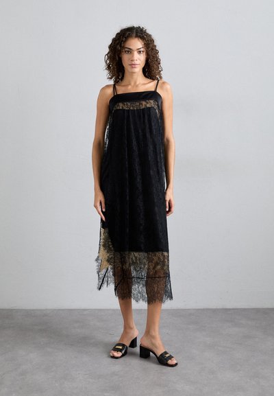 Robe en dentelle noire avec un superposé transparent, des bretelles ajustables et un ourlet en courbes. Associée à des chaussures noires à talons épais et à accents brillants.