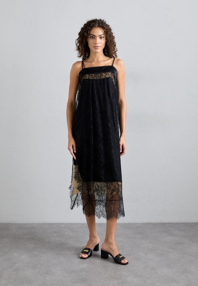 Μαύρο φόρεμα δαντέλας με διάφανη επικάλυψη, ρυθμιζόμενα λουριά και κοφτή άκρη. Συνδυασμένο με μαύρα παπούτσια με χοντρό τακούνι και γυαλιστερές λεπτομέρειες.