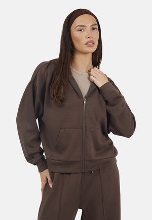 Woman wearing a brown zip-up hoodie and matching pants, with a beige top underneath and a brown headband, posing against a plain background.