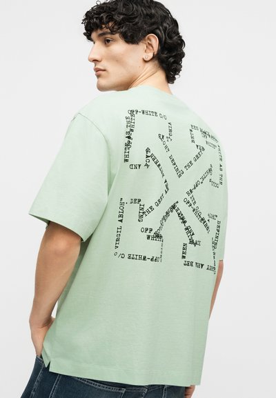 Jeune homme aux cheveux bouclés portant un t-shirt oversize vert menthe avec un texte noir formant un motif abstrait dans le dos.