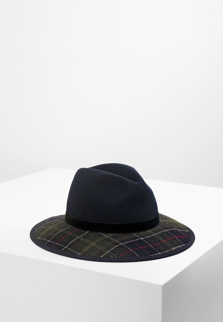 Chapeau fedora noir avec une bordure à motif à carreaux en vert foncé, rouge et blanc, posé sur un piédestal blanc sur un fond uni.