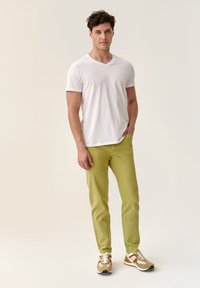 Tricou alb cu decolteu în V, pantaloni verzi skinny și adidași maro cu accente albe și portocalii, stând pe un fond neutru.