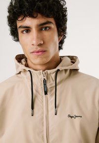 Šviesiai ruda striukė su gobtuvu, užtrauktuku priekyje, juodomis raištelėmis ir siuvinėtu "Pepe Jeans" logotipu krūtinėje. Lygi, lengva medžiaga.
