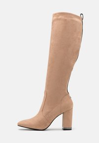 Botte beige en faux suède, hauteur genou, avec un bout pointu et un talon bloc. Présente une fermeture éclair sur le côté et une texture lisse.