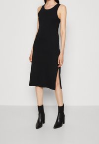 Robe noire sans manches en tissu doux, avec un design mi-long et des fentes latérales, associée à des bottines à talons noires.