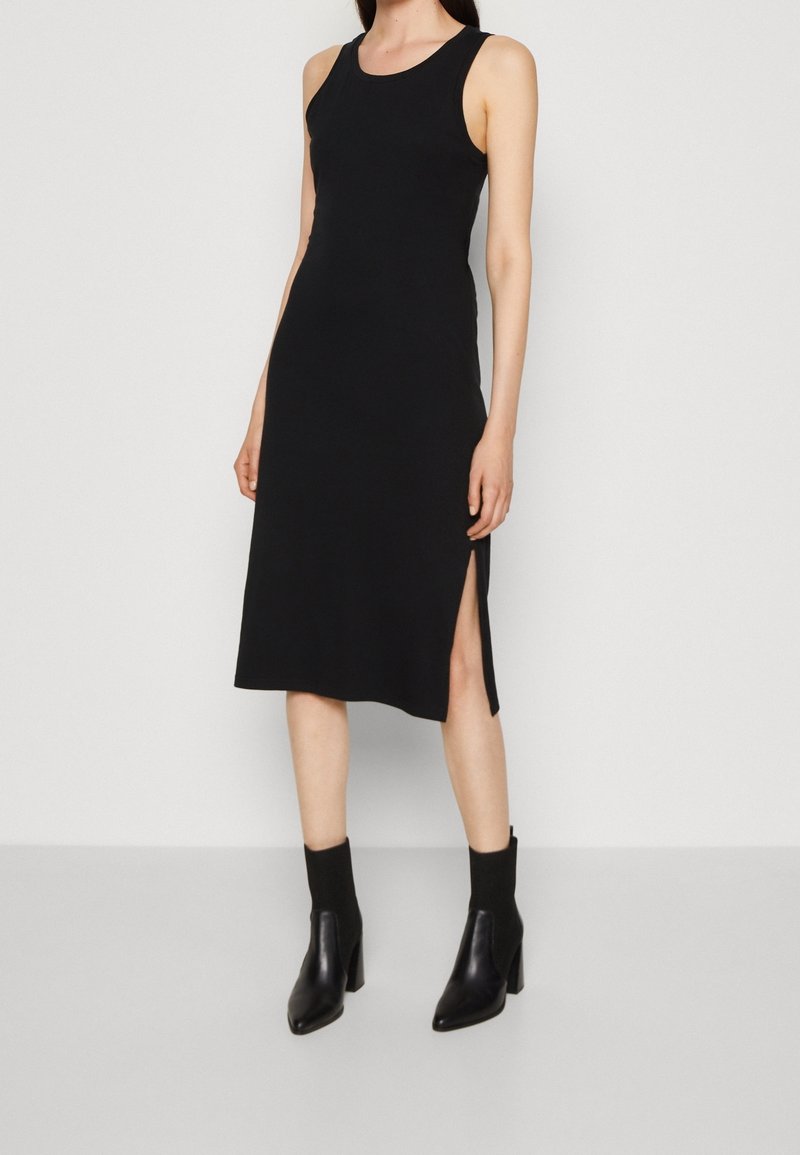 Robe noire sans manches en tissu doux, avec un design mi-long et des fentes latérales, associée à des bottines à talons noires.