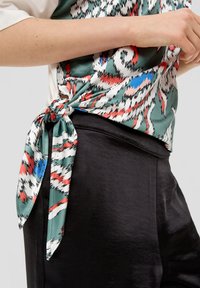 Zijde blouse in groen met abstracte patronen in rood, wit en blauw, geknoopt in de taille. Gladde zwarte stof van de broek met een subtiele glans.