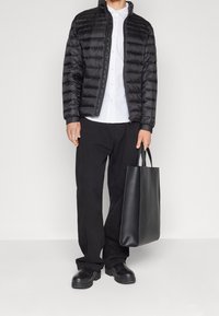 Muž v černé péřové bundě přes bílou košili, černých kalhotách a černých botách, držící černou koženou tašku za ucha.