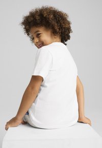 Camiseta de algodón blanca con mangas cortas, con un escote redondeado. Niño con cabello rizado sentado en un cubo blanco, mirando ligeramente hacia atrás.