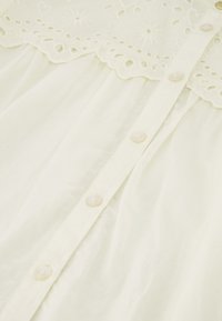 Blusa blanca con un yugo texturizado y bordado con diseños florales, suave al tacto y botones en la parte delantera.