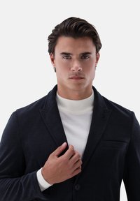 Marineblauer Cord-Blazer mit Kerbkragen, einer Brusttasche und Knopfverschluss, kombiniert mit einem weißen Rollkragenpullover. Glatte Textur.