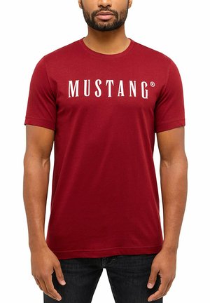 Man wearing a plain red short-sleeve T-shirt with the word "MUSTANG" printed in large white letters across the chest.