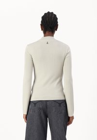 Haut à manches longues gris clair côtelé avec une coupe ajustée et un col montant. Présente un petit logo sur l'encolure au dos. Porté avec un jean foncé.