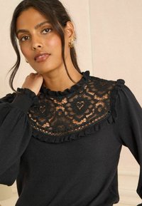 Fekete pulóver csipkebetéttel, virágmintás fodros nyakkialakítással. Bő ujjakkal és puha tapintású anyaggal.