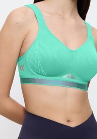 Lys grøn sports-bh med tekstureret materiale, V-hals design, mesh detaljer og et reflekterende bånd nederst.