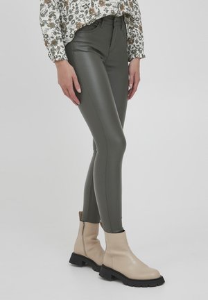 Vrouw draagt hooggetailleerde olijfgroene leren broek, bloemrijke long sleeve top en beige enkellaarzen met zwarte zolen, staand tegen een witte achtergrond.