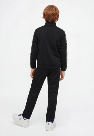 Kind mit roten Haaren steht, trägt einen schwarzen Trainingsanzug mit Logos auf den Ärmeln und weiße Sneakers mit blauen Details vor weißem Hintergrund.