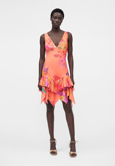 Γυναίκα που φοράει κοραλί αμάνικο φόρεμα με V λαιμόκοψη, λουλουδάτα σχέδια και ασύμμετρο βολάν στο τελείωμα, συνδυασμένο με γυμνά σανδάλια ψηλοτάκουνα.