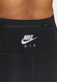 Detailní záběr na černé legíny Nike Air s malou kapsou na zip na zadním pásku, které nosí osoba s viditelnou pokožkou na střední části zad.