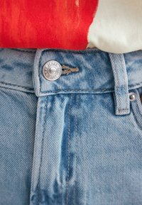 Jeans din denim de un albastru deschis prezintă un buton argintiu cu inscripția "NEW JEANS 1982" embosată, având un design clasic cu cinci buzunare și o ușoară decolorare.