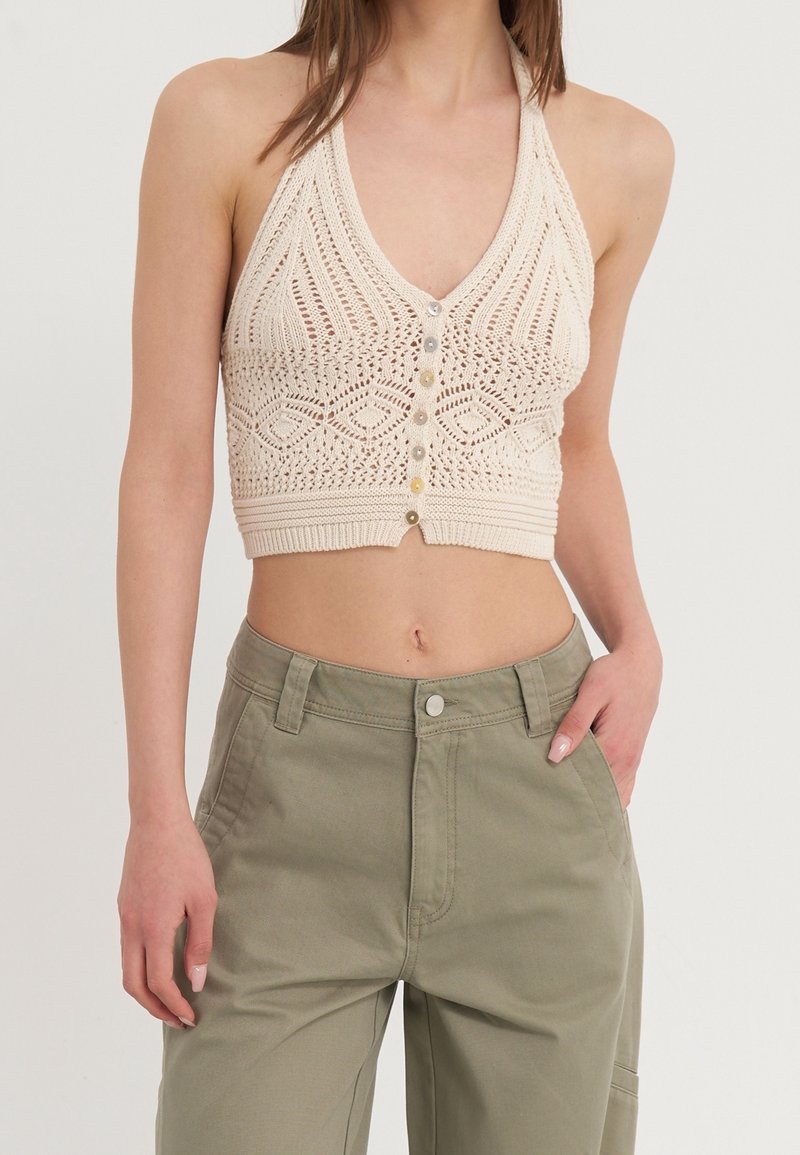 Femme portant un top court beige en crochet sans manches avec des boutons sur le devant et un pantalon taille haute vert olive, une main dans la poche.