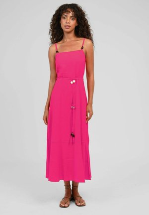 Femme aux cheveux bouclés porte une robe sans manches rose vif avec une fine ceinture et des sandales, debout devant un fond uni gris clair.