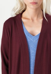 Bordeaux gebreide cardigan met een open voorkant, met een gladde textuur. Draag hem over een lichtblauwe, zachte v-hals trui.