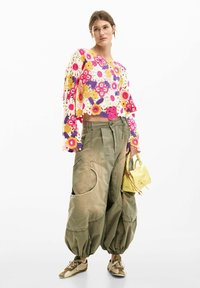 Top croșetat cu modele florale în roz, galben și mov; pantaloni cargo de culoare verde măsliniu; geantă mică galbenă; adidași de culoare bej.