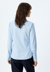 Camisa de manga larga azul claro con cuello de solapa, puños abotonados y un diseño ajustado. Tela suave con un ligero brillo.