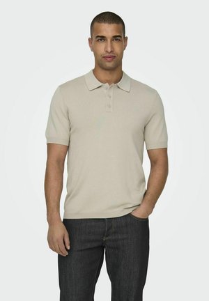Camisa polo beige de suave tejido de punto, con un cuello clásico, tapeta de tres botones y mangas cortas, combinada con jeans de mezclilla oscura.