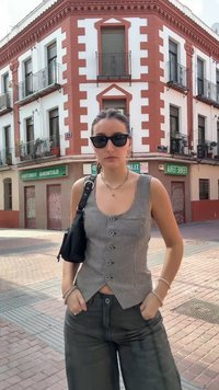 Γυναίκα που φορά γυαλιά ηλίου, καρό αμάνικο τοπ και σκούρο παντελόνι, στέκεται σε μια γωνία δρόμου της πόλης με ένα κόκκινο και λευκό κτίριο στο φόντο.