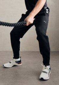 Persoon in zwarte Puma-sweatpants en witte Puma-sneakers die een dikke zwarte battle rope vasthoudt en aan het trainen is op een grijze vloer.