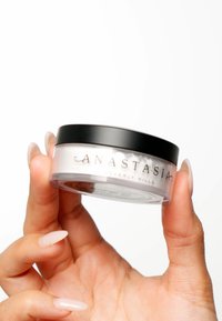 Transparante pot met een zwart deksel, gelabeld "Anastasia Beverly Hills." De pot bevat een witte poeder en wordt vastgehouden door een hand met lange, bleke nagels.