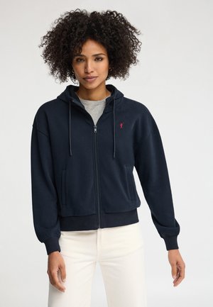 Woman with curly hair wearing a navy zip-up hoodie over a gray shirt and white pants, standing against a plain light background.