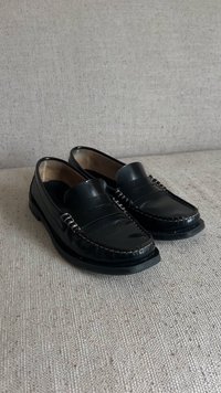 Black leather loafers with a smooth finish, featuring a stitched accent around the toe and a rounded profile. Flat rubber sole.