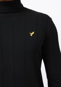 Suéter negro de cuello alto acanalado con un emblema de pájaro bordado en dorado en el lado izquierdo del pecho. La tela parece suave y con textura.