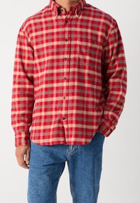 Czerwona i kremowa koszula flanelowa w kratę z kołnierzykiem zapinanym na guziki, długimi rękawami, kieszenią na piersi, zaokrąglonym dołem i czarnymi guzikami. Połączona z niebieskimi dżinsami.