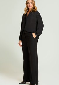 Blouse noire avec accents bijoutés sur les épaules et les poignets, associée à un pantalon noir taille haute présentant un design aux lignes épurées.