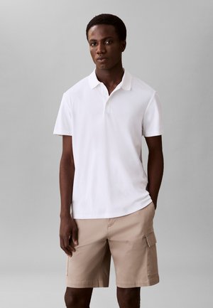 Bărbat tânăr purtând un tricou polo alb cu mâneci scurte și pantaloni scurți cargo bej, stând în picioare pe un fundal simplu gri.