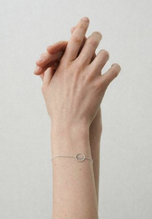 Dwie splecione dłonie z delikatną srebrną bransoletką z zawieszką w kształcie rozbłysku słońca na nadgarstku na jednolitym tle.