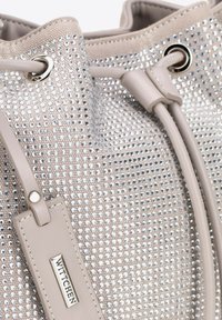 Sac seau en beige texturé avec des sequins, des sangles ajustables et une étiquette logo. Des œillets métalliques accentuent la fermeture à cordon.