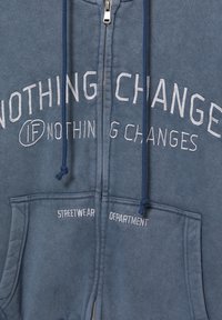 Zip-Hoodie aus verblichem blauen Baumwollstoff. Mit weißem, gesticktem Text und Kordelzügen. Zwei Vorderaschen mit kontrastierenden gestickten Etiketten.