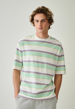 Junger Mann mit lockigem Haar, der ein pastellgestreiftes kurzärmeliges Hemd und hellgraue Hose trägt, steht vor einer schlichten hellen Wand.