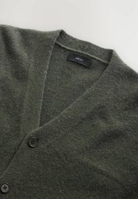 Grön ullcardigan med V-hals, två mörka knappar och en texturerad yta. Lappen visar "NEXT."