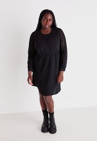 Robe noire avec un design à manches longues en dentelle, taille froncée et coupe décontractée. Associée à des bottes à plateforme noires épaisses. Texture lisse.