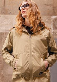 Bež zip-up jopica z elastičnimi manšeti in stranskimi žepi, ki ima majhen črn logotip. Model nosi sončna očala na teksturirani podlagi.